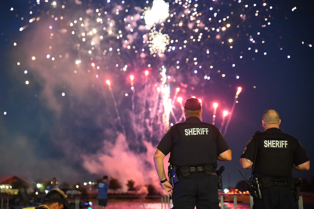 Dois xerifes ficam de costas para a câmara, a observar fogos de artifício coloridos a explodir no céu noturno acima de uma multidão junto à água — uma atmosfera tão fiável e impressionante como o topo do WordPress Hosting.